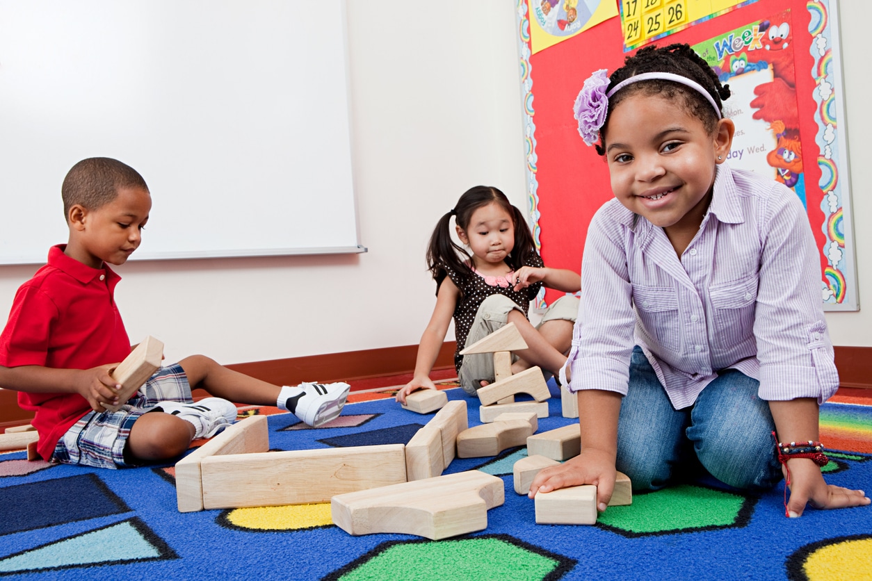 Three children sit on a rug playing together with building blocks
