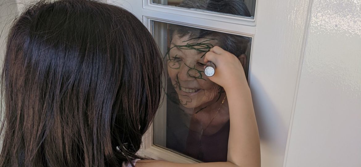 A young girl draws on a window over a woman's face standing on the other side of the window