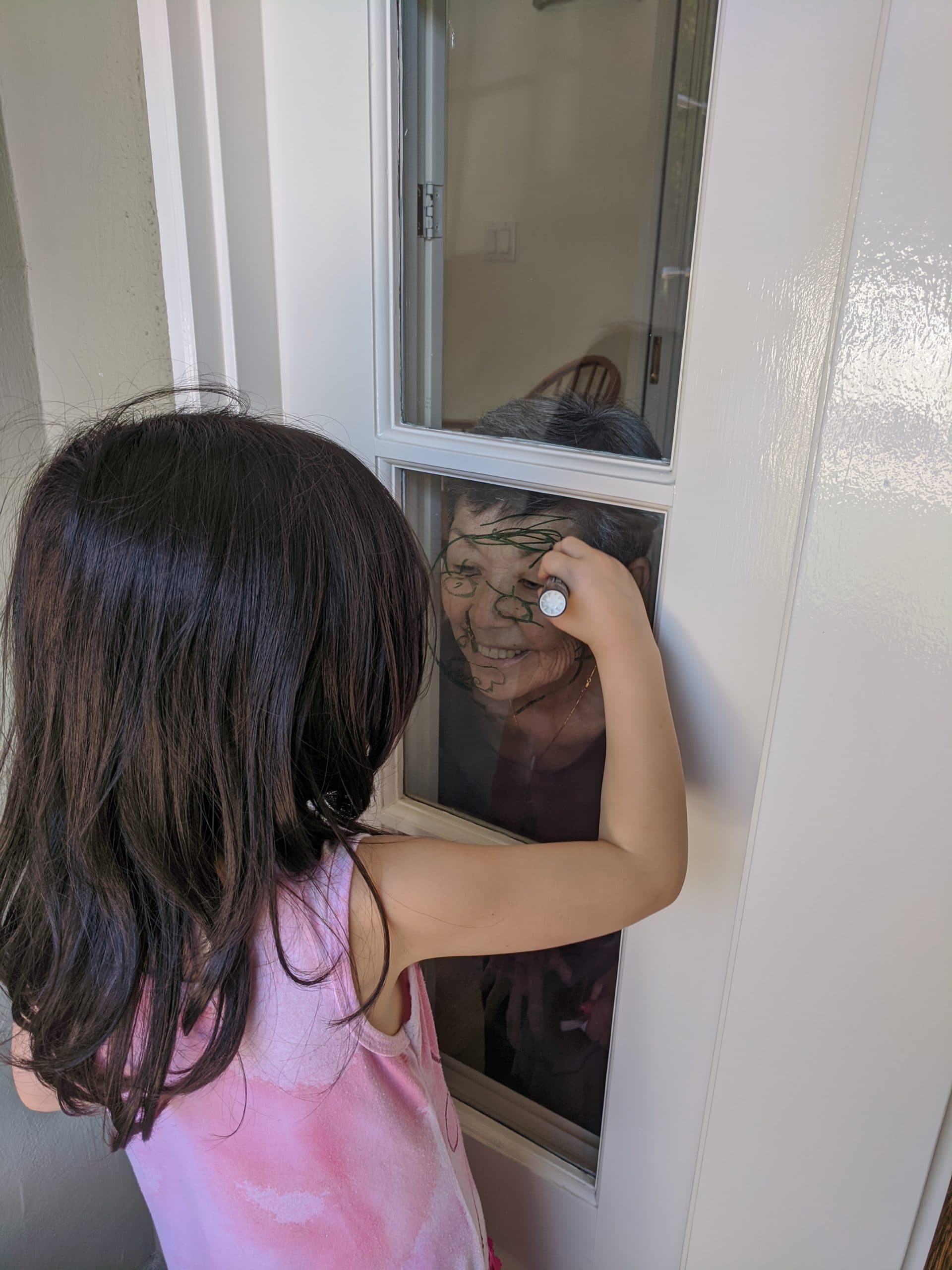 A young girl draws on a window over a woman's face standing on the other side of the window