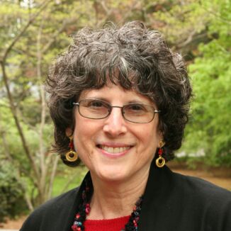 A smiling woman with curly hair, deeply involved in early childhood education, wearing glasses, earrings, and a necklace outdoors.
