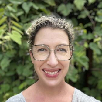 Woman with curly hair and glasses, specializing in early childhood education, smiling in front of a leafy background.