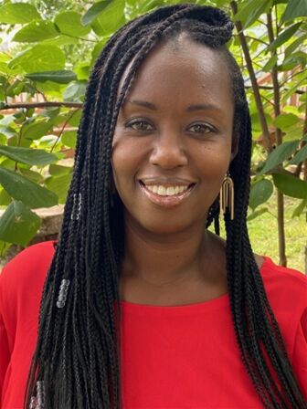 A smiling woman with braided hair, wearing a red top and earrings, standing in front of green foliage. A smiling woman with braided hair, wearing a red top and earrings, standing in front of green foliage.