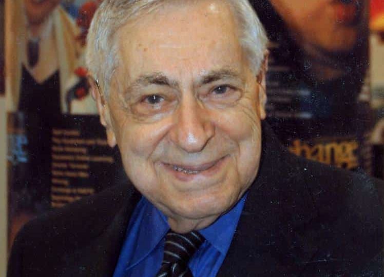 An elderly man with a friendly expression wearing a blue shirt and blazer at an indoor event.