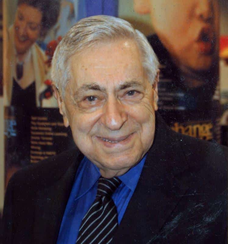 An elderly man with a friendly expression wearing a blue shirt and blazer at an indoor event.