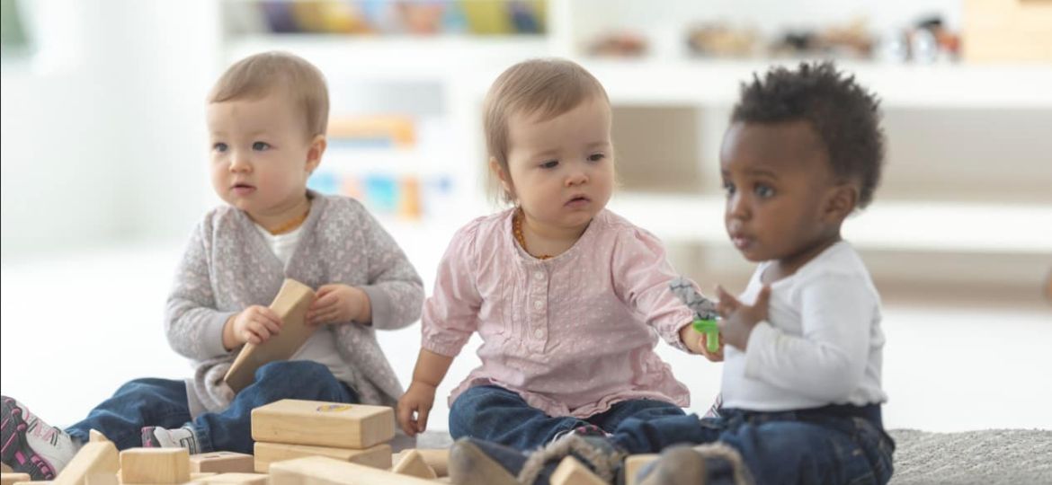 Three toddlers play with wooden blocks on a rug, surrounded by scattered toys in a bright playroom.