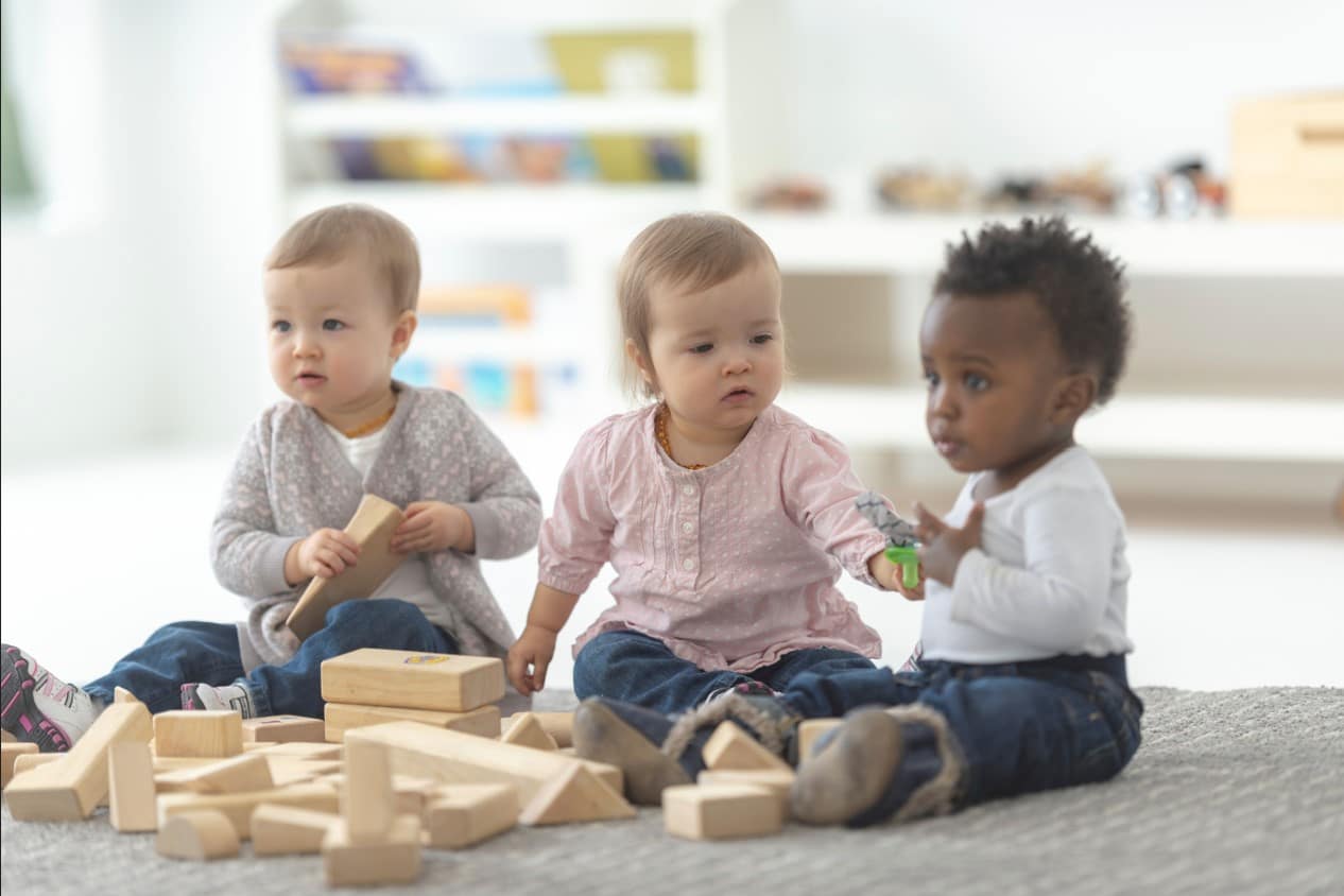 Three toddlers play with wooden blocks on a rug, surrounded by scattered toys in a bright playroom.