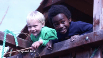 Two young children, one with blond hair and one with curly black hair, smile and lean over a wooden railing on an elevated wooden structure. Text appears in the image, capturing a moment reminiscent of early childhood videos.