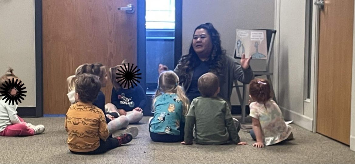A woman is seated on the floor in front of a small group of young children, reading a book to them in a classroom setting.