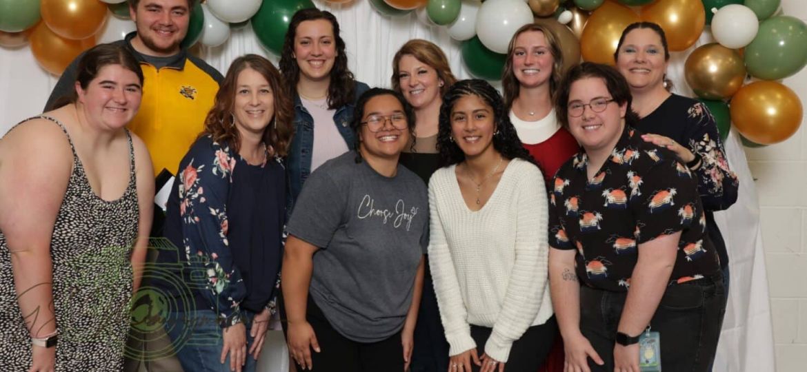 A group of people posing together in front of a balloon garland with green, white, and gold balloons.