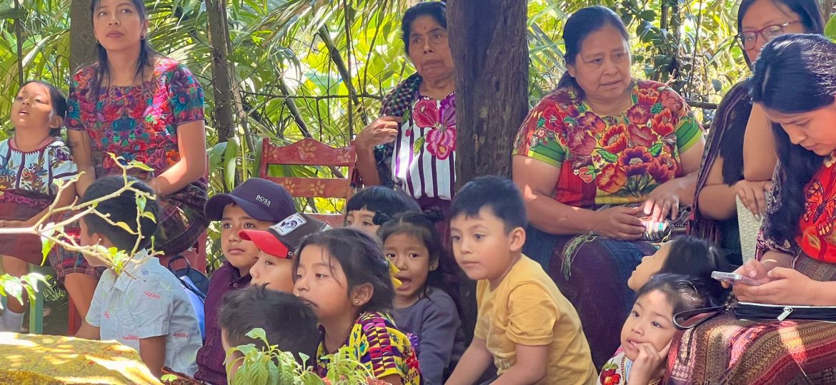 A group of people, including children and women in colorful clothing, sit outdoors surrounded by plants and trees. Some are seated while others crouch on the ground.