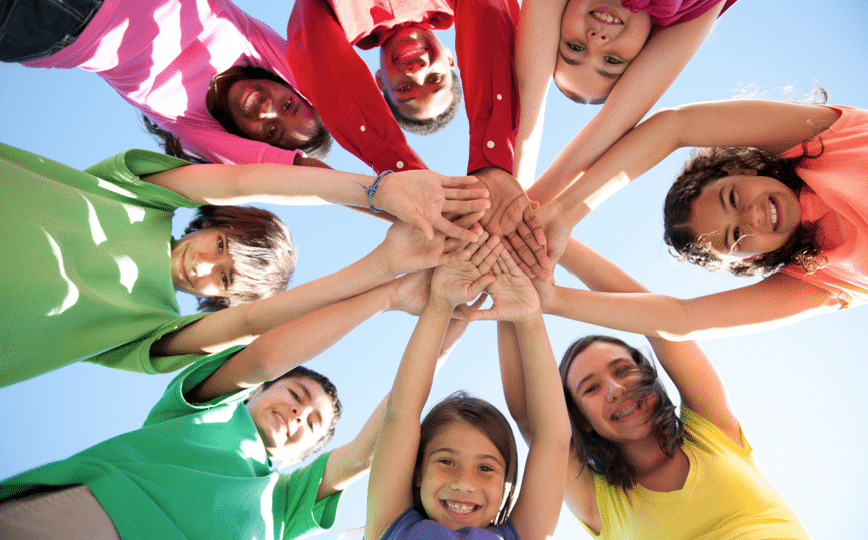 A group of nine diverse children stand in a circle, reaching in to place their hands together in the center, viewed from below against a clear blue sky.
