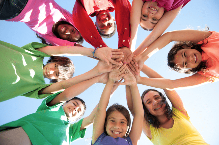 A group of nine diverse children stand in a circle, reaching in to place their hands together in the center, viewed from below against a clear blue sky.