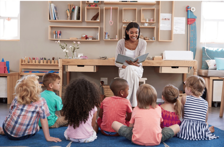 A teacher reads a book to a group of attentive young children seated on the floor in a classroom.