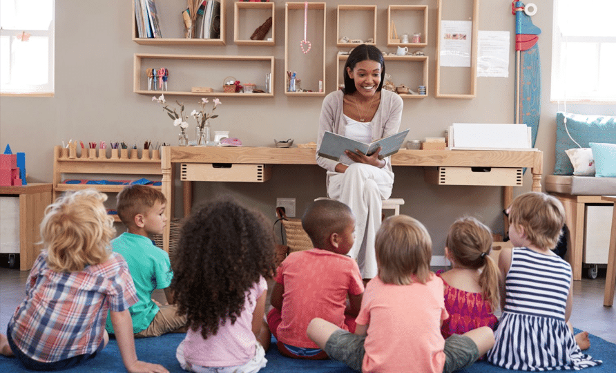 A teacher reads a book to a group of attentive young children seated on the floor in a classroom.