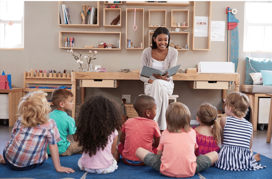 A teacher reads a book to a group of attentive young children seated on the floor in a classroom.