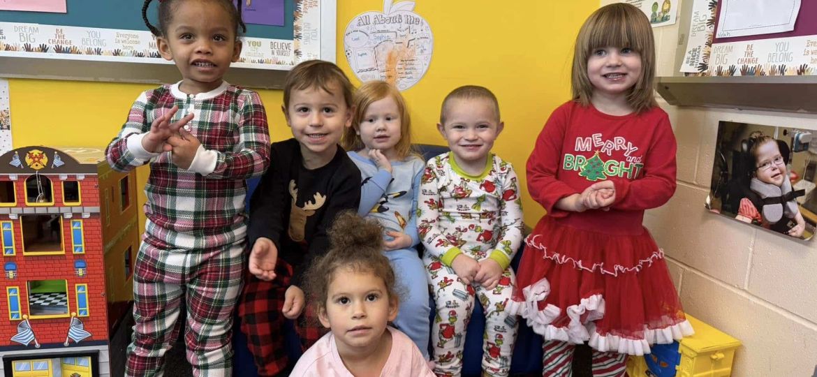 A group of six children wearing festive outfits, posing indoors in front of colorful bulletin boards and a toy house.