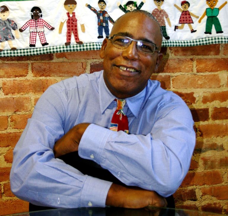 A man in glasses, wearing a blue shirt and colorful tie, sits with arms crossed on a glass table, smiling in front of a brick wall and quilt with human figures.