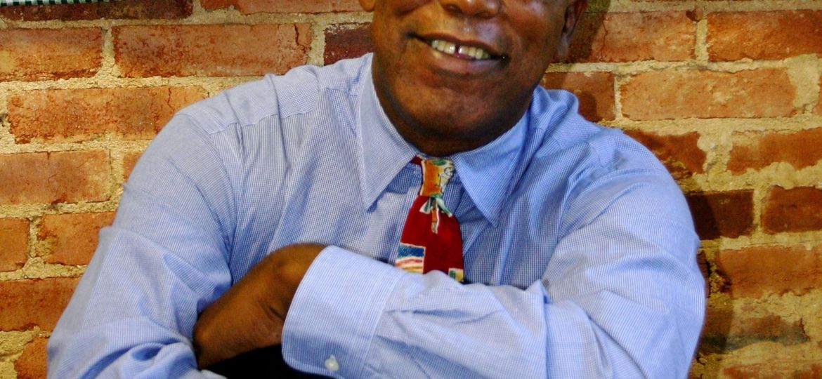 A man in glasses, wearing a blue shirt and colorful tie, sits with arms crossed on a glass table, smiling in front of a brick wall and quilt with human figures.