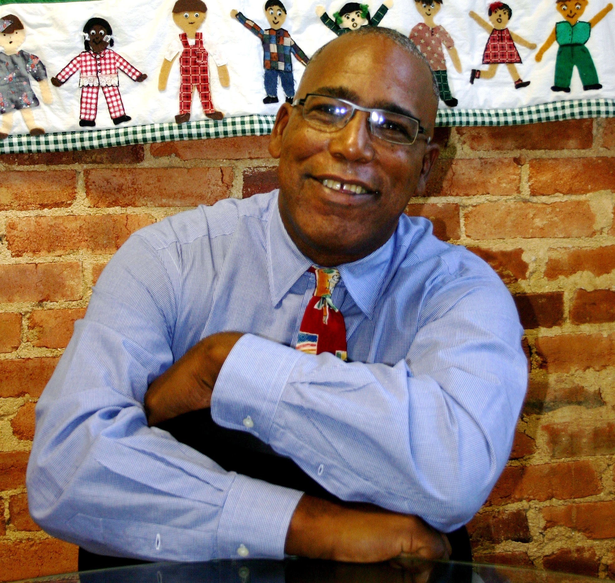 A man in glasses, wearing a blue shirt and colorful tie, sits with arms crossed on a glass table, smiling in front of a brick wall and quilt with human figures.