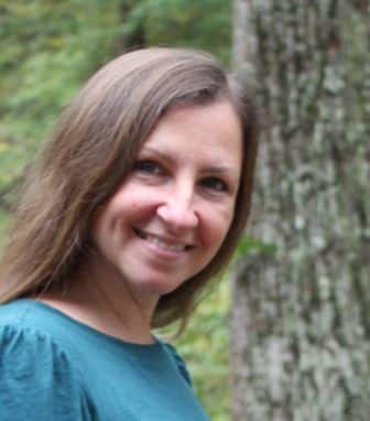 A woman with long brown hair and a teal top smiles outdoors in front of a tree and greenery. A woman with long brown hair and a teal top smiles outdoors in front of a tree and greenery.