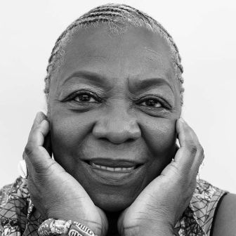 Elderly woman with braided hair smiles softly, resting her face in her hands. She wears rings and beaded bracelets. The photo is black and white with a plain background. Elderly woman with braided hair smiles softly, resting her face in her hands. She wears rings and beaded bracelets. The photo is black and white with a plain background.