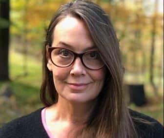 A woman with long brown hair and glasses stands outdoors, facing the camera, with blurred green and yellow foliage in the background. A woman with long brown hair and glasses stands outdoors, facing the camera, with blurred green and yellow foliage in the background.