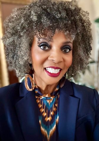 A woman with curly gray hair, wearing a navy blazer, patterned top, and large hoop earrings, smiling at the camera. A woman with curly gray hair, wearing a navy blazer, patterned top, and large hoop earrings, smiling at the camera.