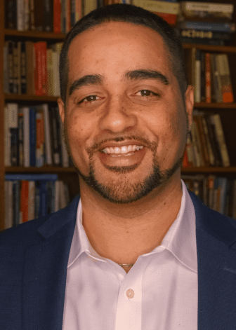 A man in a suit and collared shirt smiles in front of a bookshelf filled with books. A man in a suit and collared shirt smiles in front of a bookshelf filled with books.