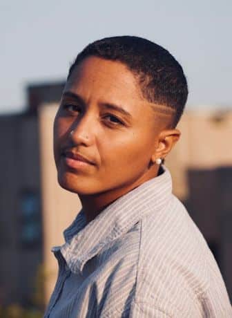 Person with short hair and two nose piercings wearing a striped button-up shirt, standing outdoors with a building in the background. Person with short hair and two nose piercings wearing a striped button-up shirt, standing outdoors with a building in the background.