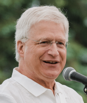 An older man with white hair and glasses speaks into a microphone outdoors, wearing a white collared shirt. An older man with white hair and glasses speaks into a microphone outdoors, wearing a white collared shirt.