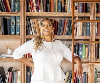 A woman in a white top stands smiling with one hand on a wooden ladder, in front of a bookshelf filled with assorted books. A woman in a white top stands smiling with one hand on a wooden ladder, in front of a bookshelf filled with assorted books.