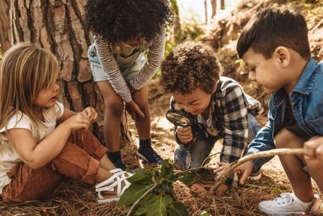 Four young children examine plants and leaves on the forest floor, with one child using a magnifying glass while the others watch closely.