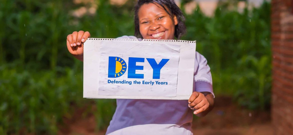 A person stands outdoors holding a sign that reads "DEY Defending the Early Years," with green foliage visible in the background.