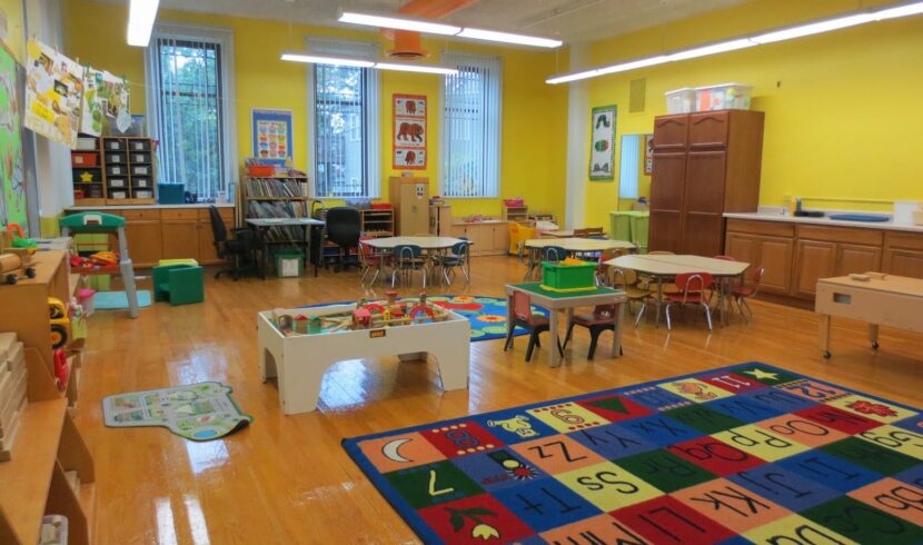 A brightly lit preschool classroom with yellow walls, small tables and chairs, educational toys, and an alphabet rug on a shiny wooden floor.