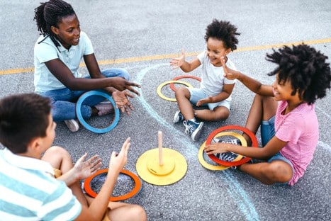 Four children sit on pavement in a circle, playing a ring toss game with colored rings and a wooden peg, smiling and interacting.